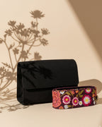 Black clutch with floral-patterned embroidered handle on a beige surface with plant shadows.