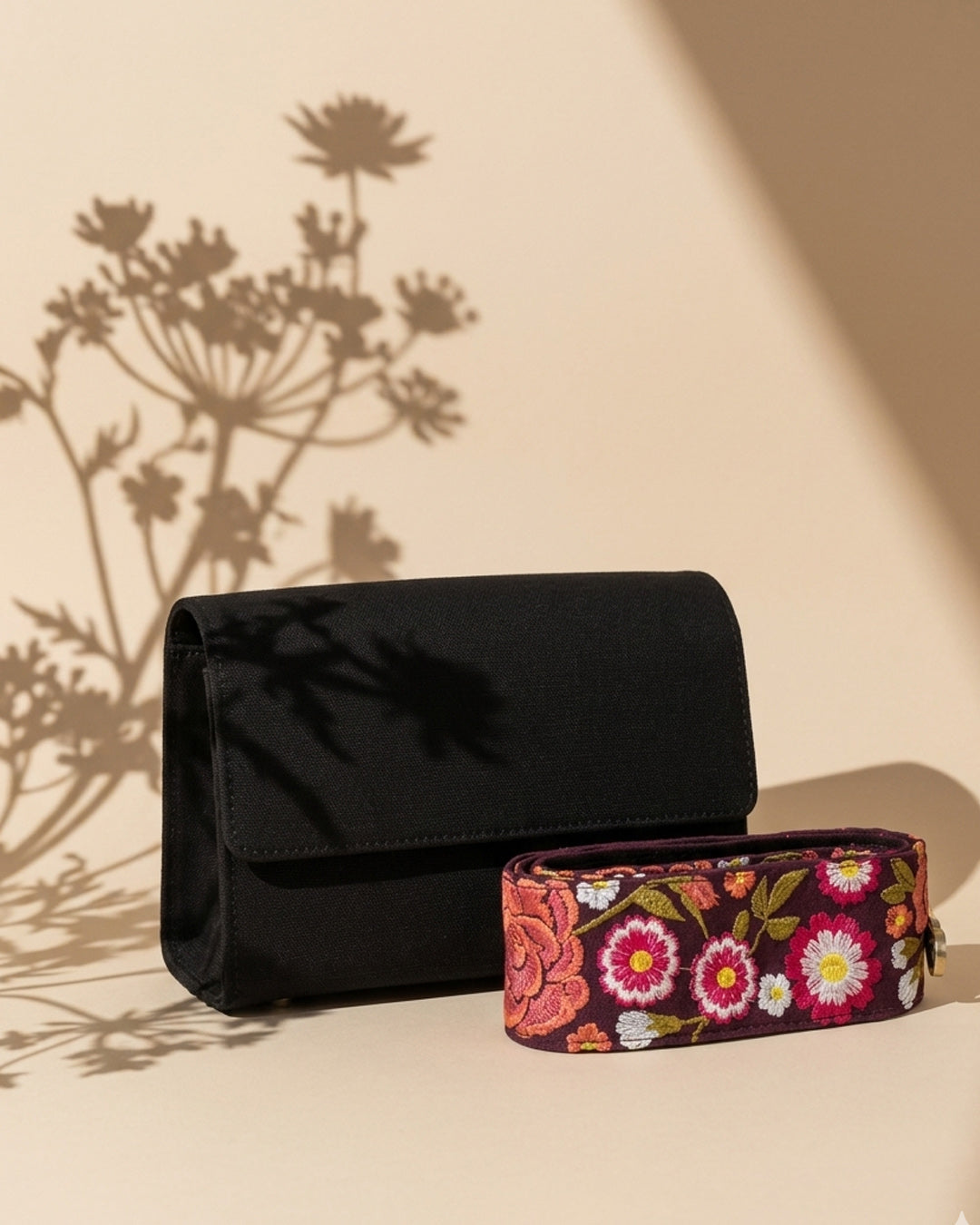 Black clutch with floral-patterned embroidered handle on a beige surface with plant shadows.