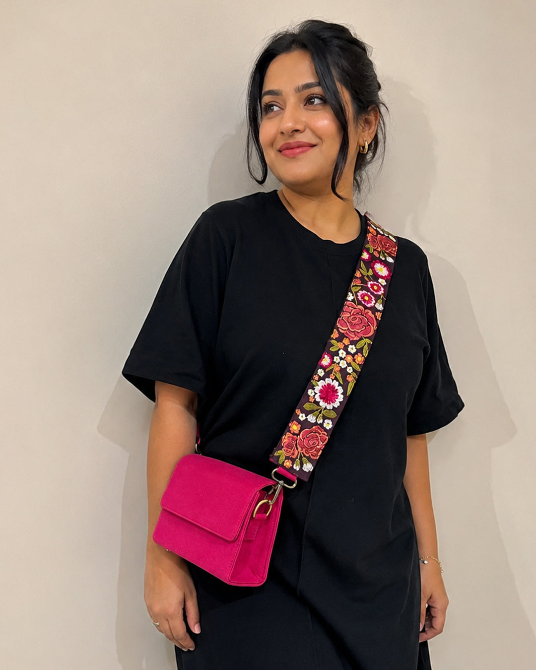 Indian woman wearing a black outfit with a pink sling bag featuring a floral embroidered strap against a plain background